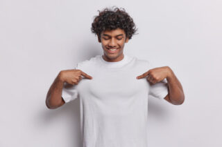 Waist up shot of cheerful curly haired Hindu man points at blank t shirt with both index fingers shows place for your design being in good mood isolated over white background. Mock up space for logo